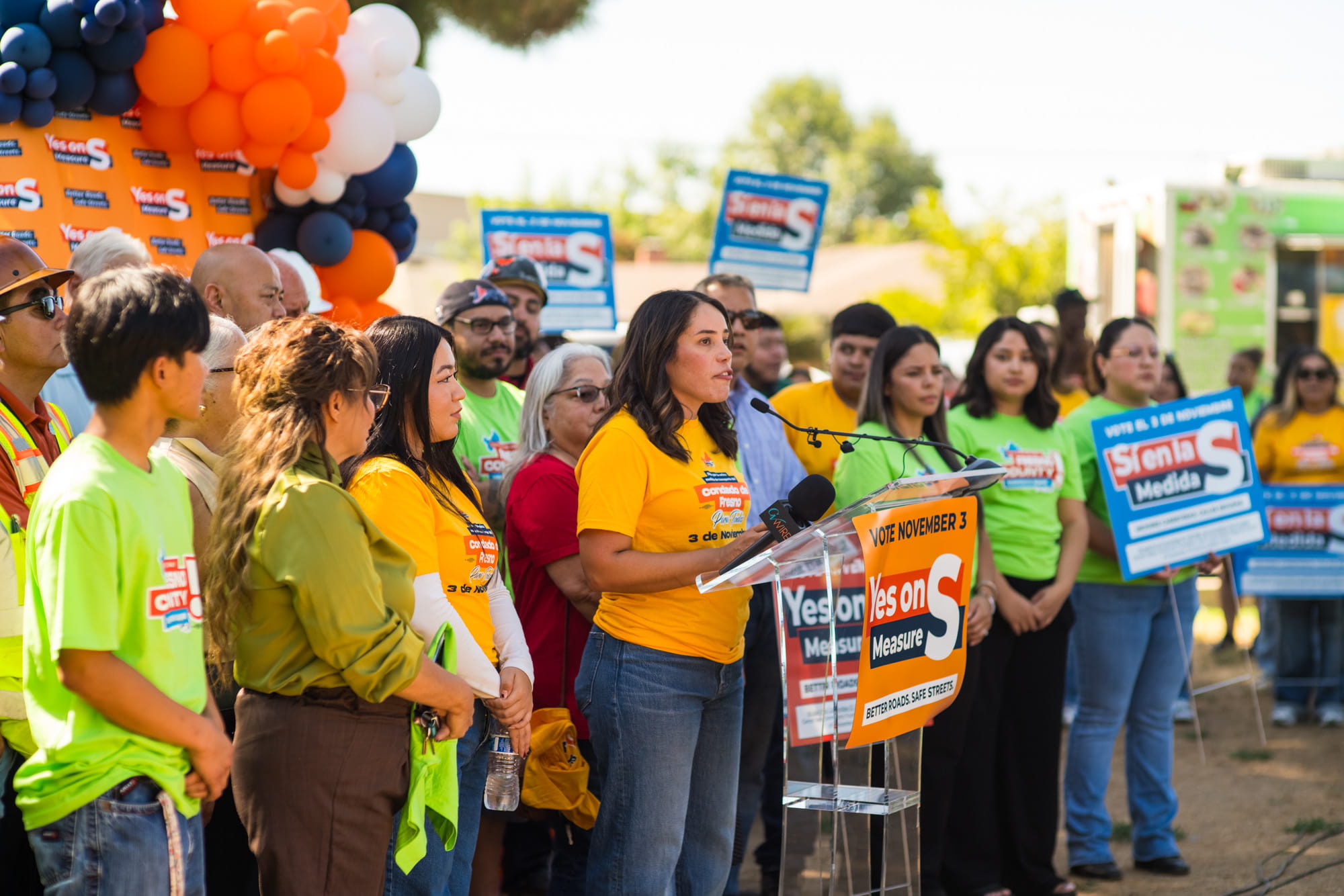 Woman speaks at a Yes on Measure S press event surrounded by youth, workers, and residents supporting modern transportation investments.